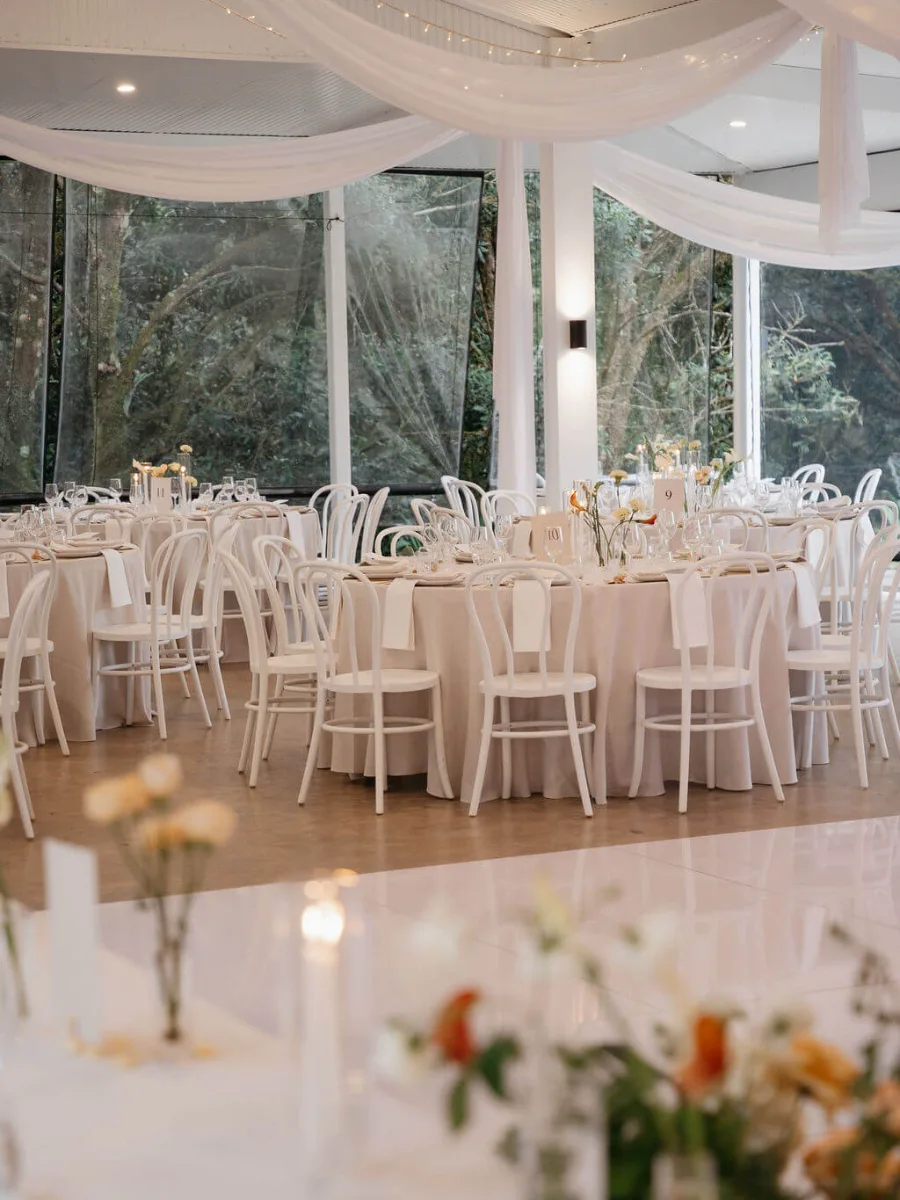 Round tables with white bentwood chairs beneath draped white fabric and fairy lights, with tall bud-vase florals in the foreground