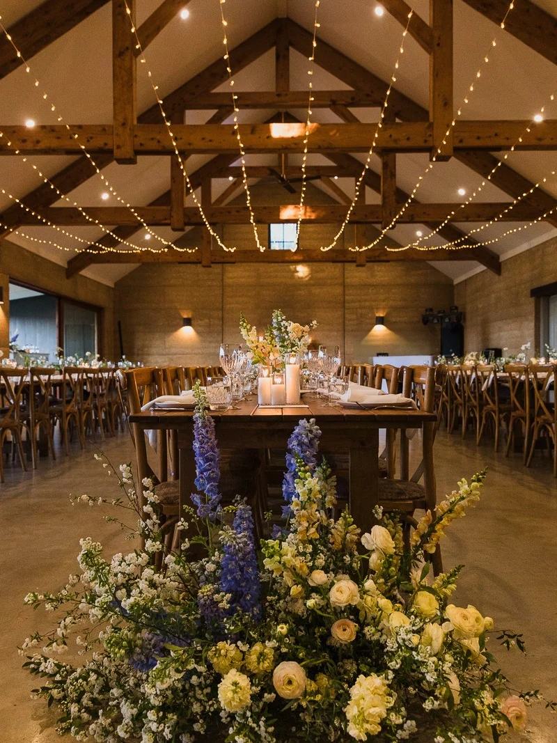 Timber head table behind a large floor arrangement of blue delphinium, white and yellow blooms, beneath exposed rafters strung with fairy lights