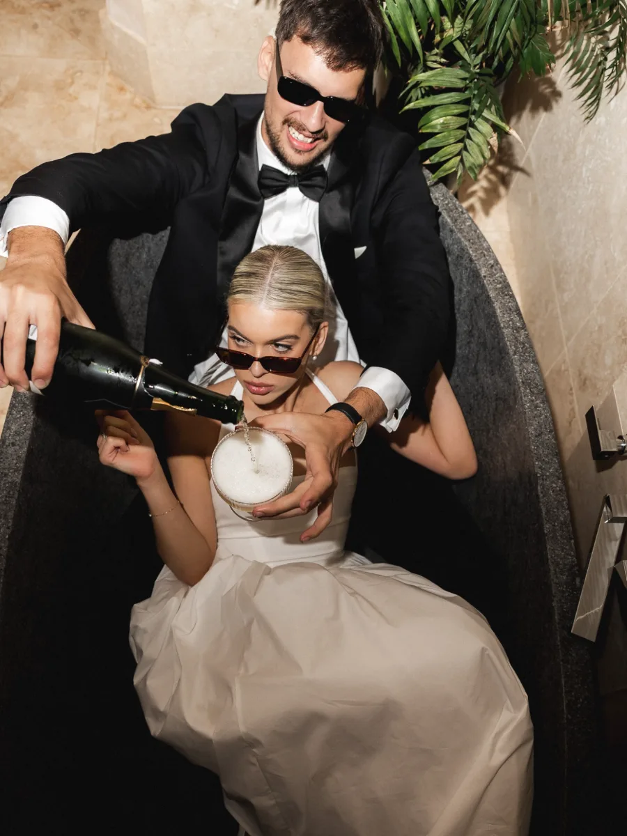 Groom in a tuxedo and sunglasses pouring champagne into a coupe held by his sunglasses-wearing bride as they sit together in a stone bath
