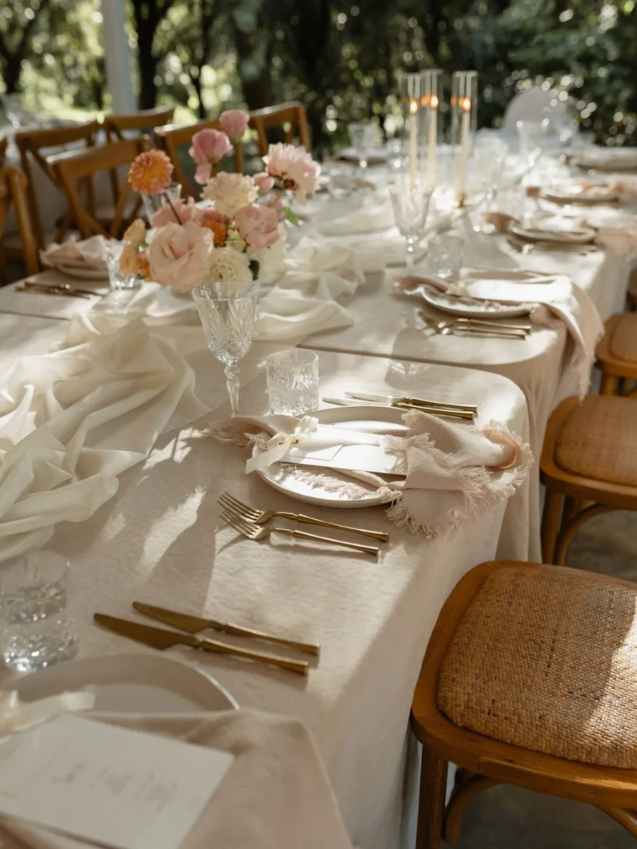 Long cream-linen table with a draped silk runner, blush and peach roses, taper candles, gold cutlery and woven cross-back chairs