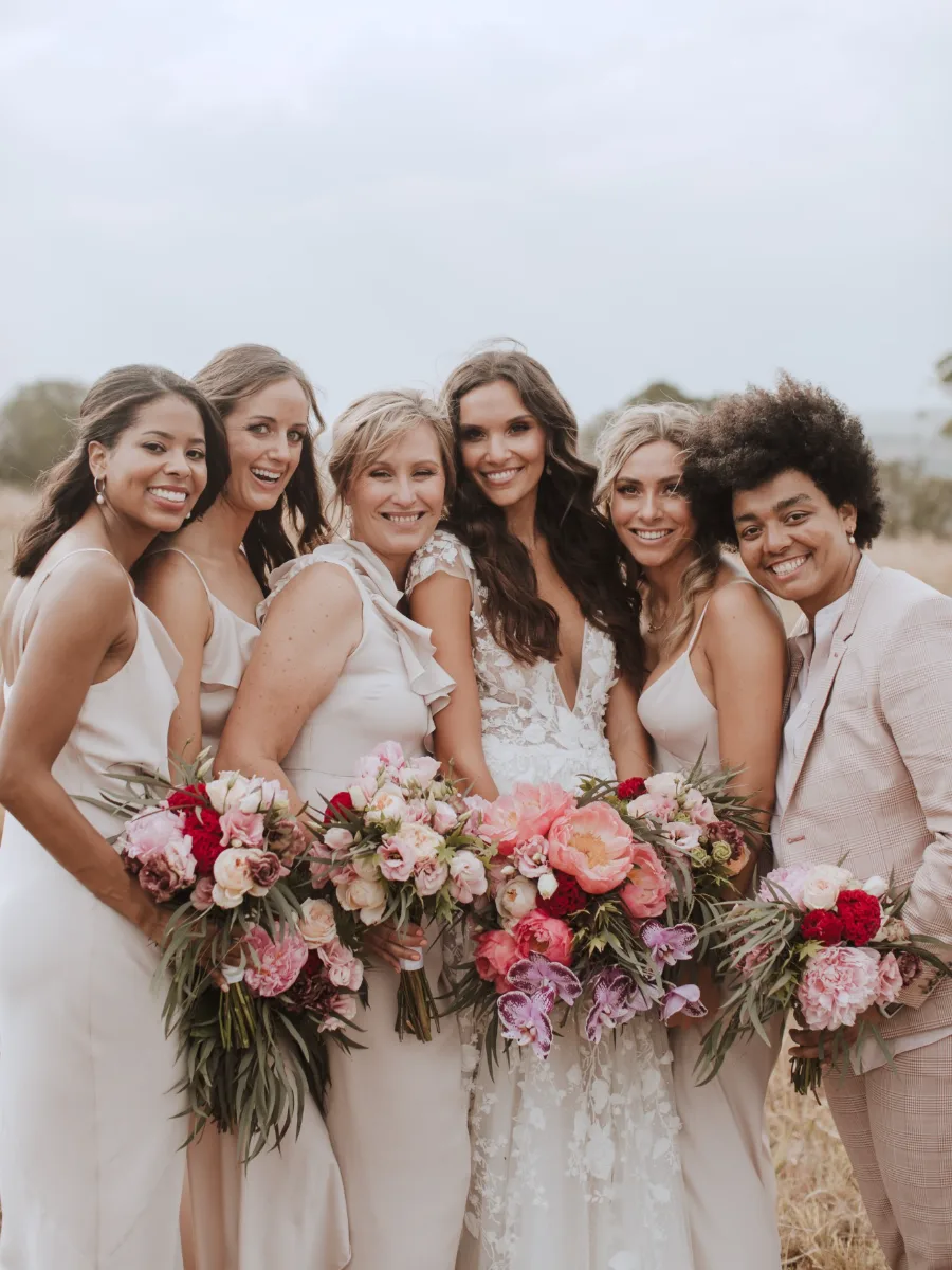 Bride and five bridesmaids smiling close together, each holding a bouquet of pink, coral and red blooms