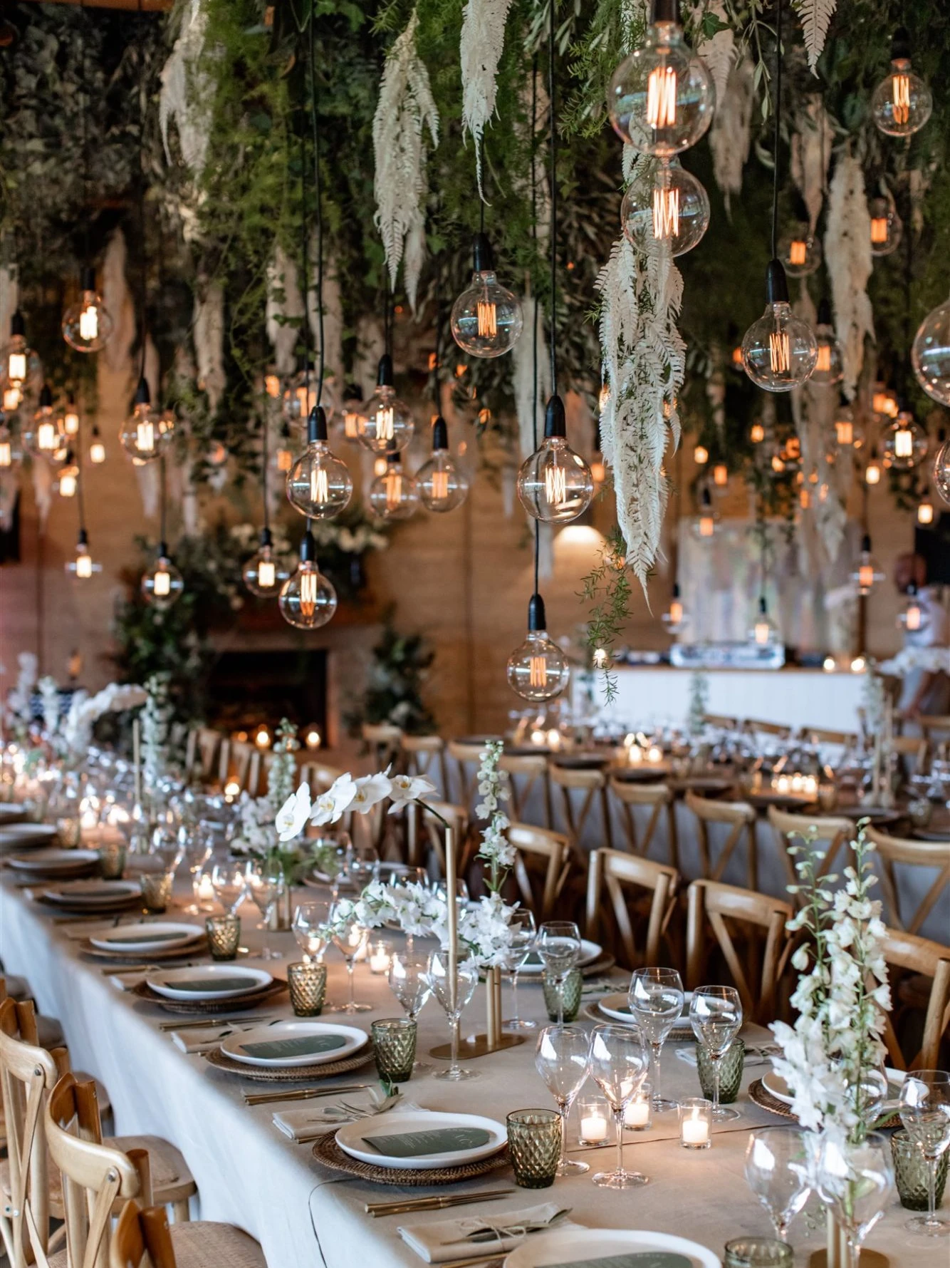 Long linen-covered reception table with candles, green glassware and white orchids beneath a canopy of greenery, white ferns and hanging filament bulbs