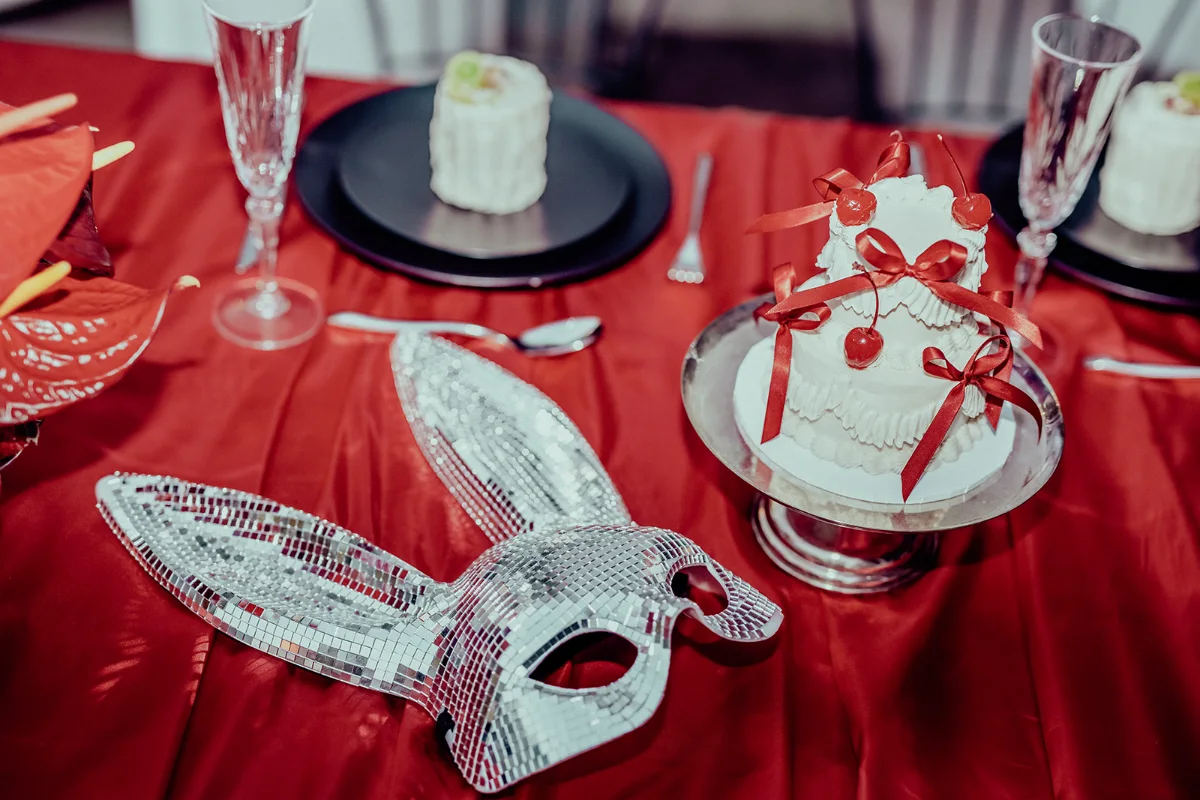 Mirrored disco bunny mask and a mini two-tier cake with red bows and cherries on a red satin tablecloth beside champagne flutes and black plates