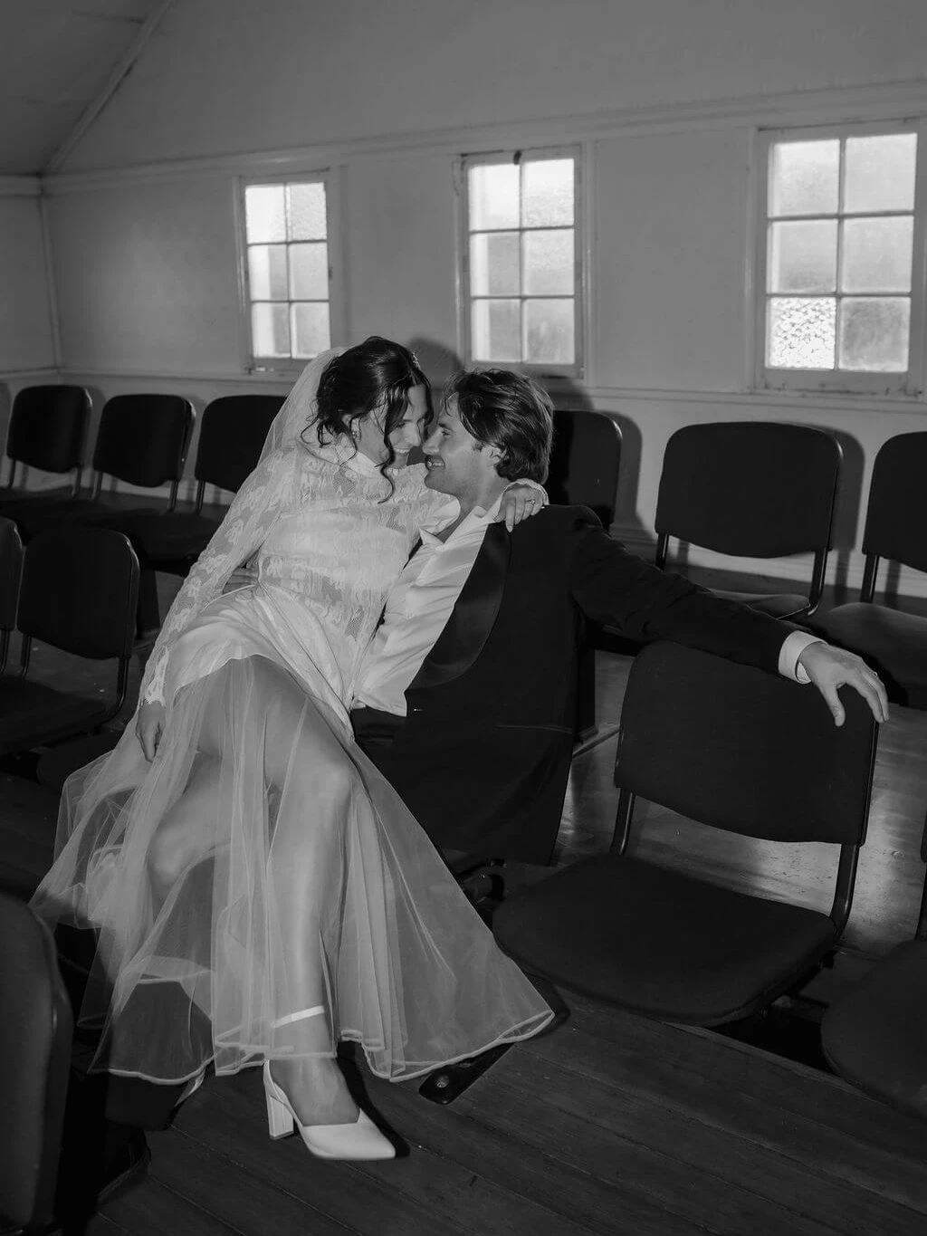 Black and white image of a smiling bride in a lace-sleeved gown and tulle skirt sitting on her groom's knee in an empty hall of black chairs, nose to nose