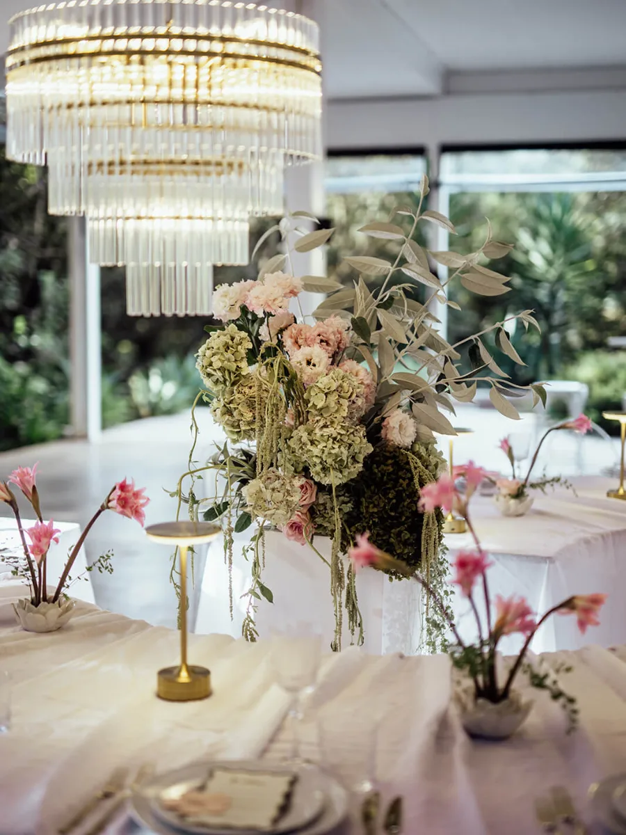 Tall pastel hydrangea and eucalyptus centrepiece under a crystal chandelier, with gold table lamps and pink amaryllis in small vases