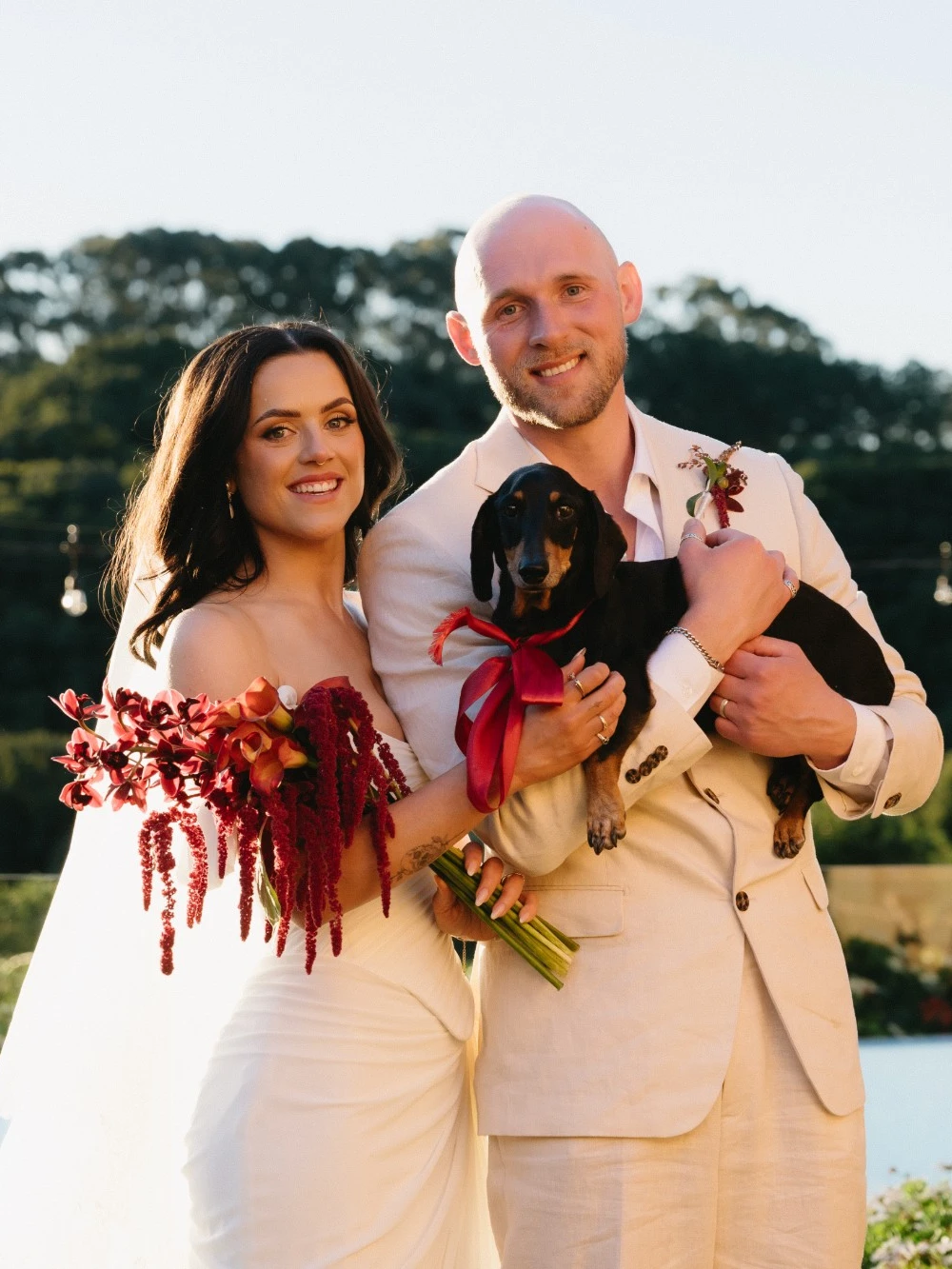 Smiling bride with a deep red bouquet and groom in a linen suit holding a dachshund with a red ribbon, above an infinity pool