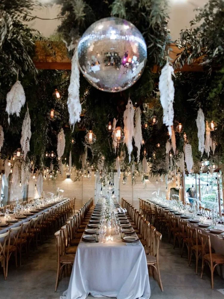 Long banquet table beneath a mirrored disco ball, hanging white feathers, pendant bulbs and a canopy of greenery
