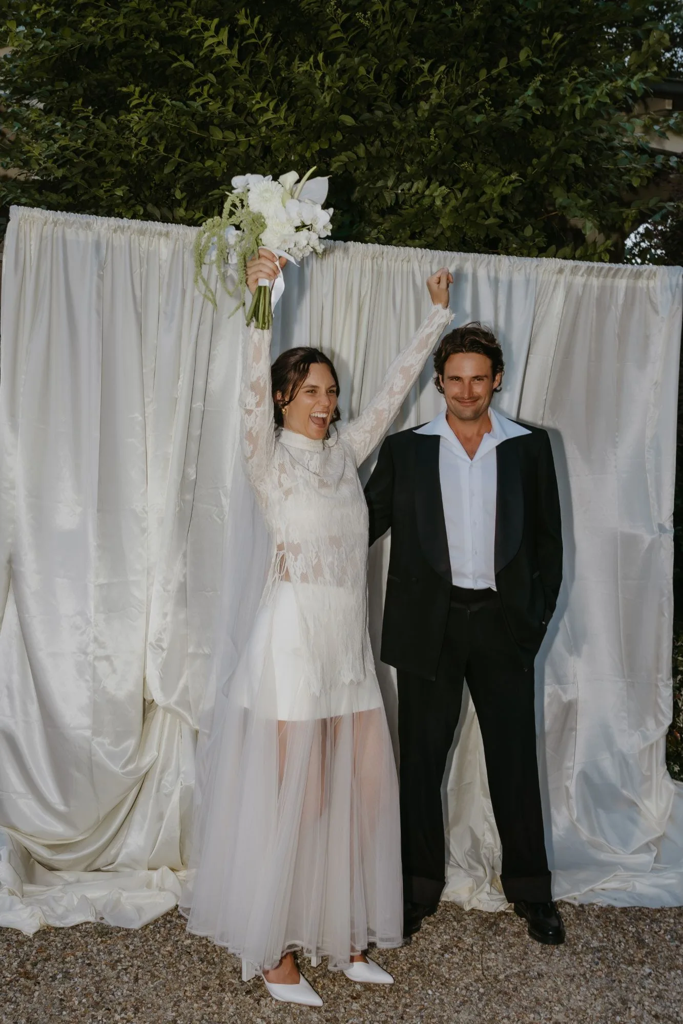 Bride cheering with a white bouquet raised overhead beside her groom in a black tuxedo, in front of cream satin drapes and a hedge