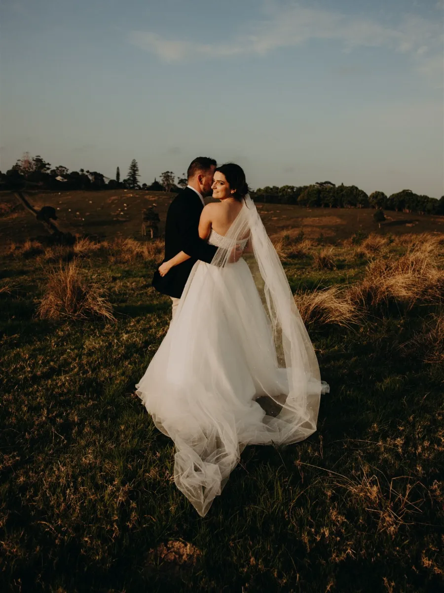 Groom whispering to his smiling bride as she looks back over her shoulder, her tulle skirt and pearl-dotted veil trailing across a golden paddock at dusk