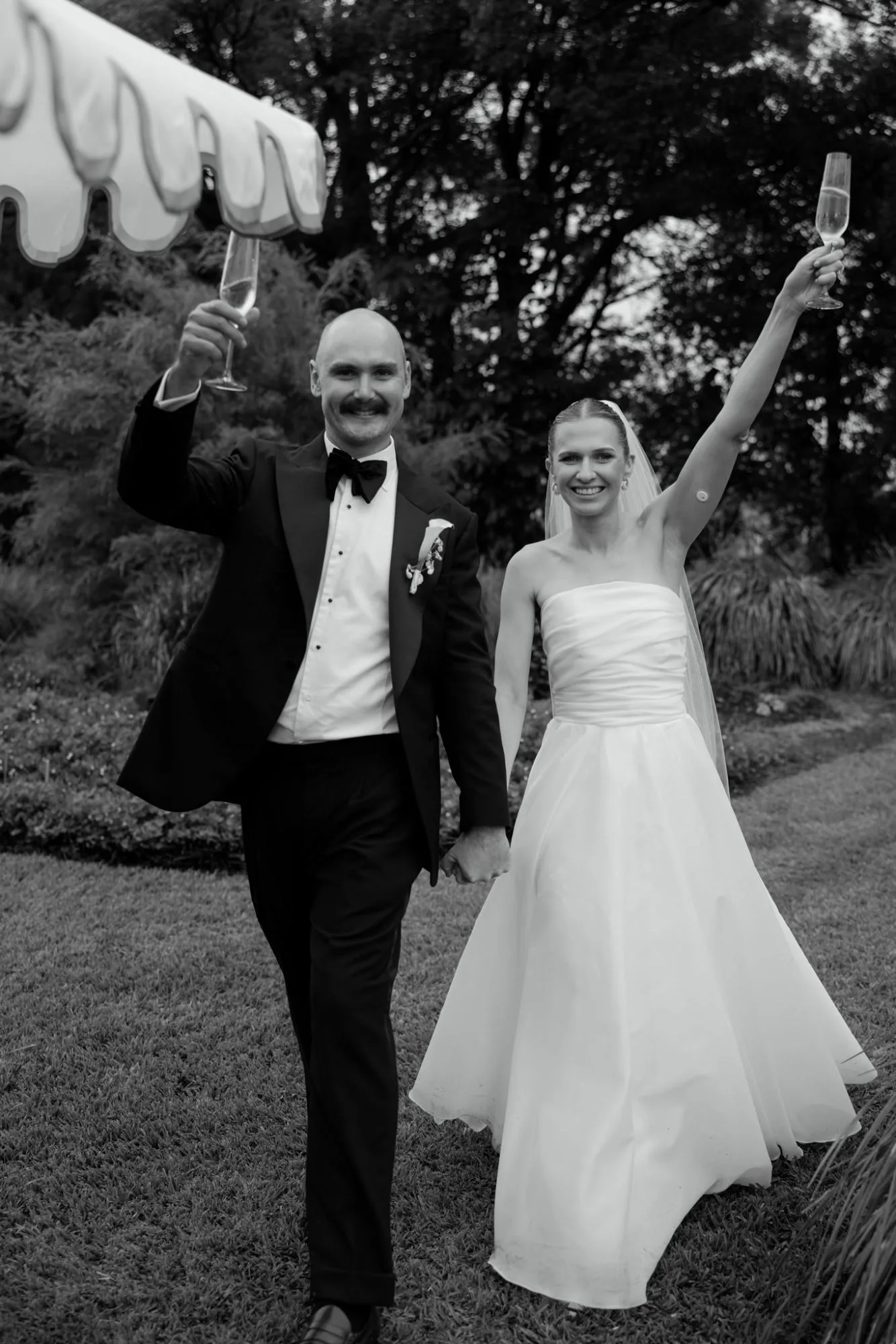 Black and white image of a smiling couple walking hand in hand with champagne flutes raised, a scalloped umbrella at the edge of the frame