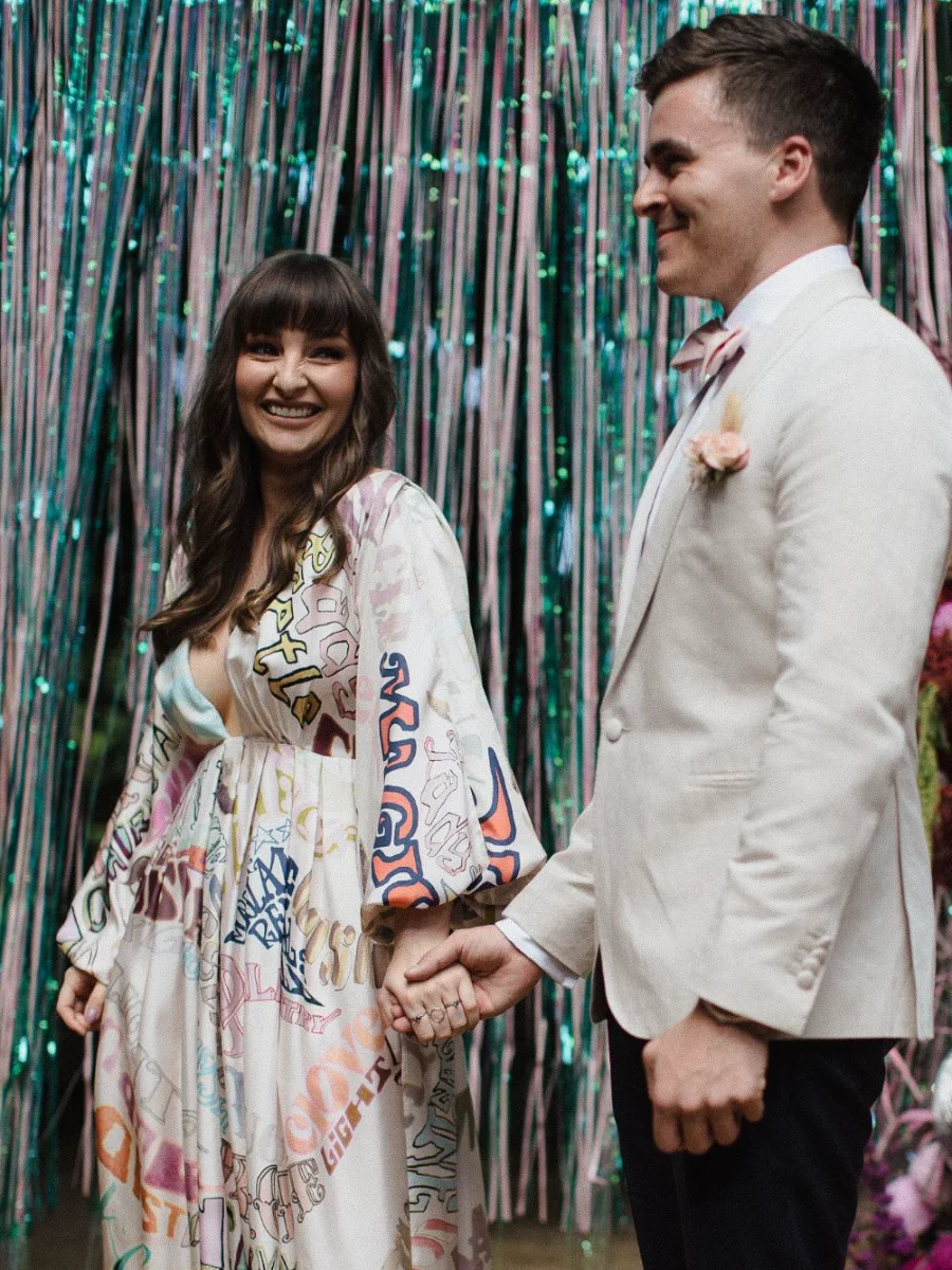 Smiling bride in a hand-lettered graffiti-print gown holding hands with her groom in a cream jacket in front of an iridescent tinsel curtain