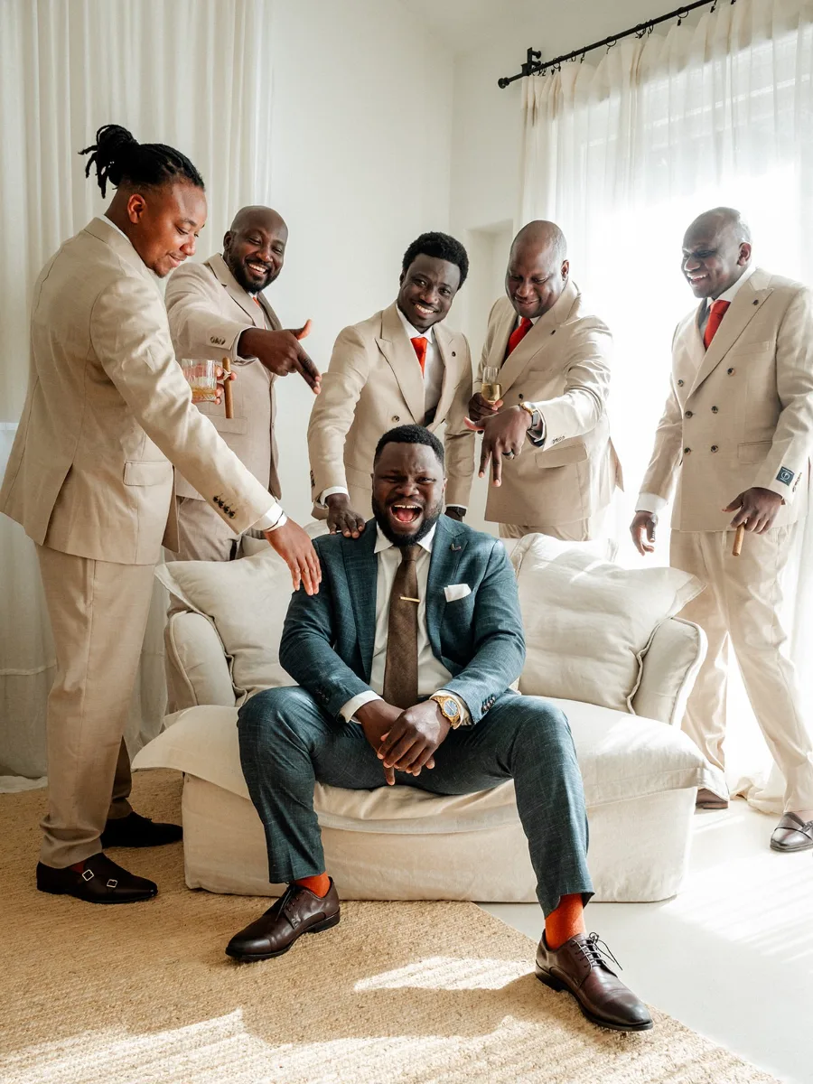 Groomsmen in sand-coloured suits laughing and gesturing at the seated groom in a bright, airy room