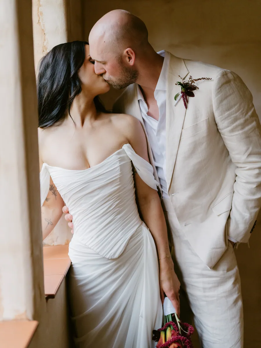 Couple sharing a kiss beside a window, bride holding a deep red bouquet