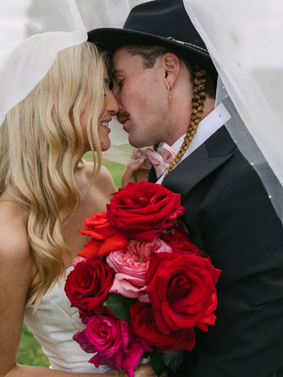 Bride and groom in a black hat touching noses under a pearl-dotted veil, a bouquet of red and pink roses between them