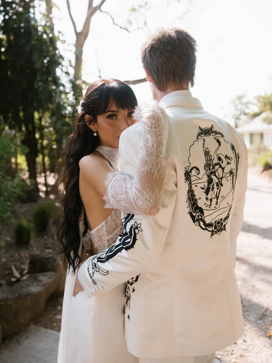 Bride in a lace-sleeved gown glancing over the shoulder of her groom, whose white jacket is embroidered with a rodeo scene
