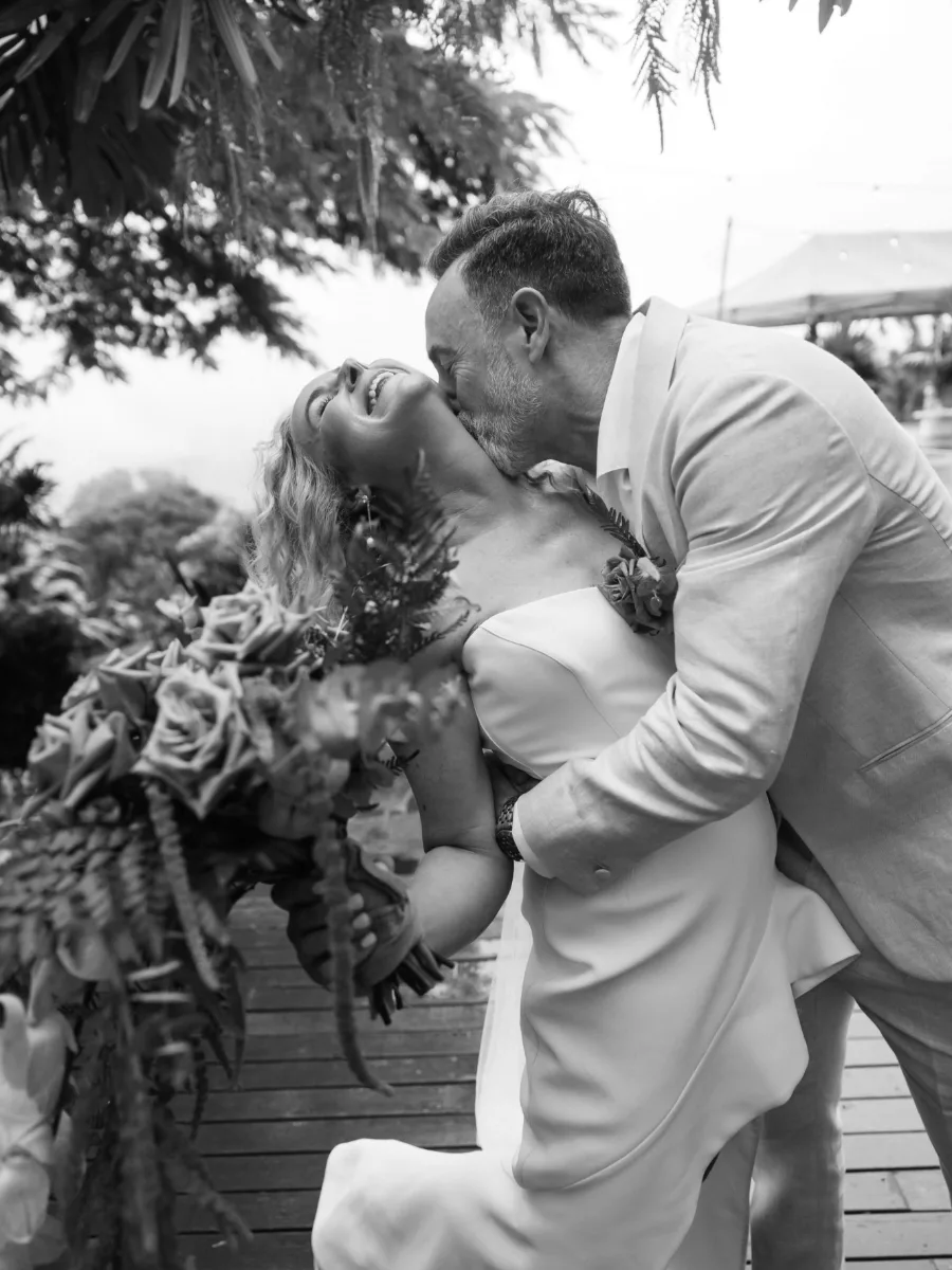 Black and white image of a laughing bride dipped back as her groom kisses her neck, her bouquet in hand