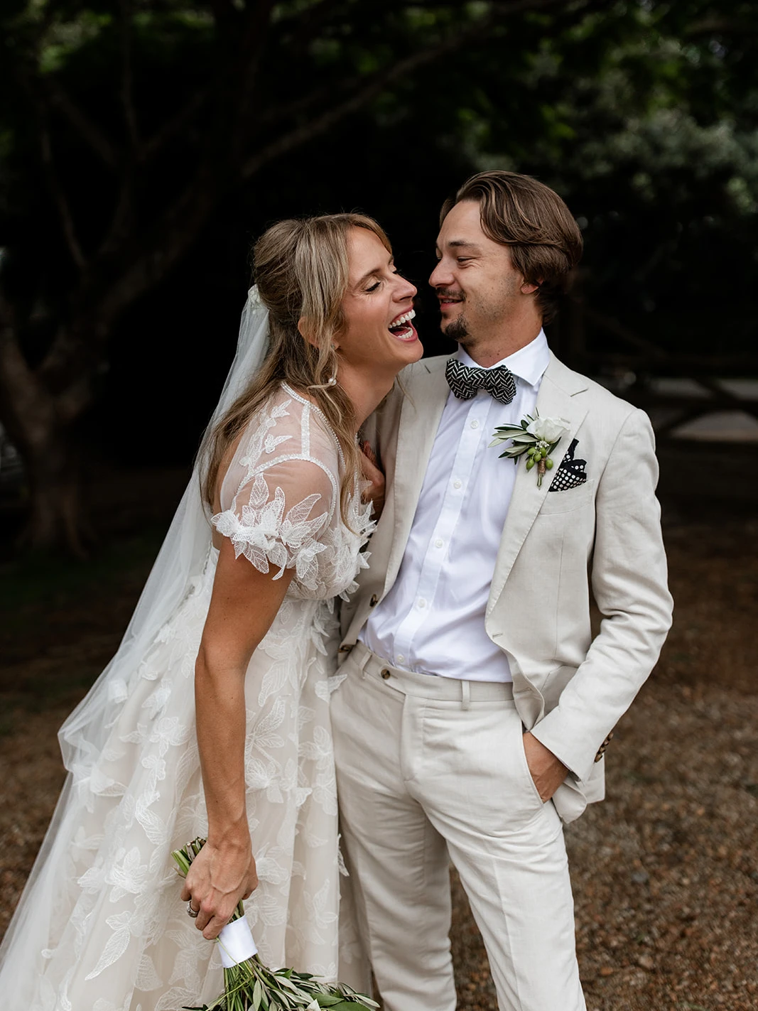 Laughing bride in a leaf-lace gown and veil leaning into her groom in a cream linen suit and black-and-white bow tie