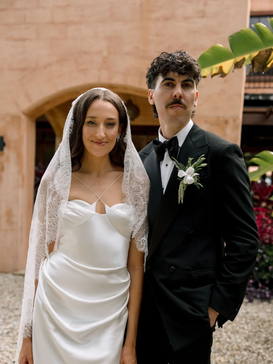Bride in a lace mantilla veil and satin halter gown standing beside her groom in a black tuxedo in front of a terracotta archway