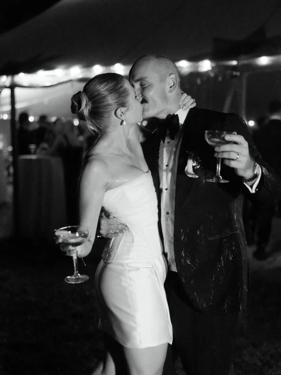 Black and white image of a couple kissing at night beneath a marquee strung with festoon lights, each holding a coupe glass