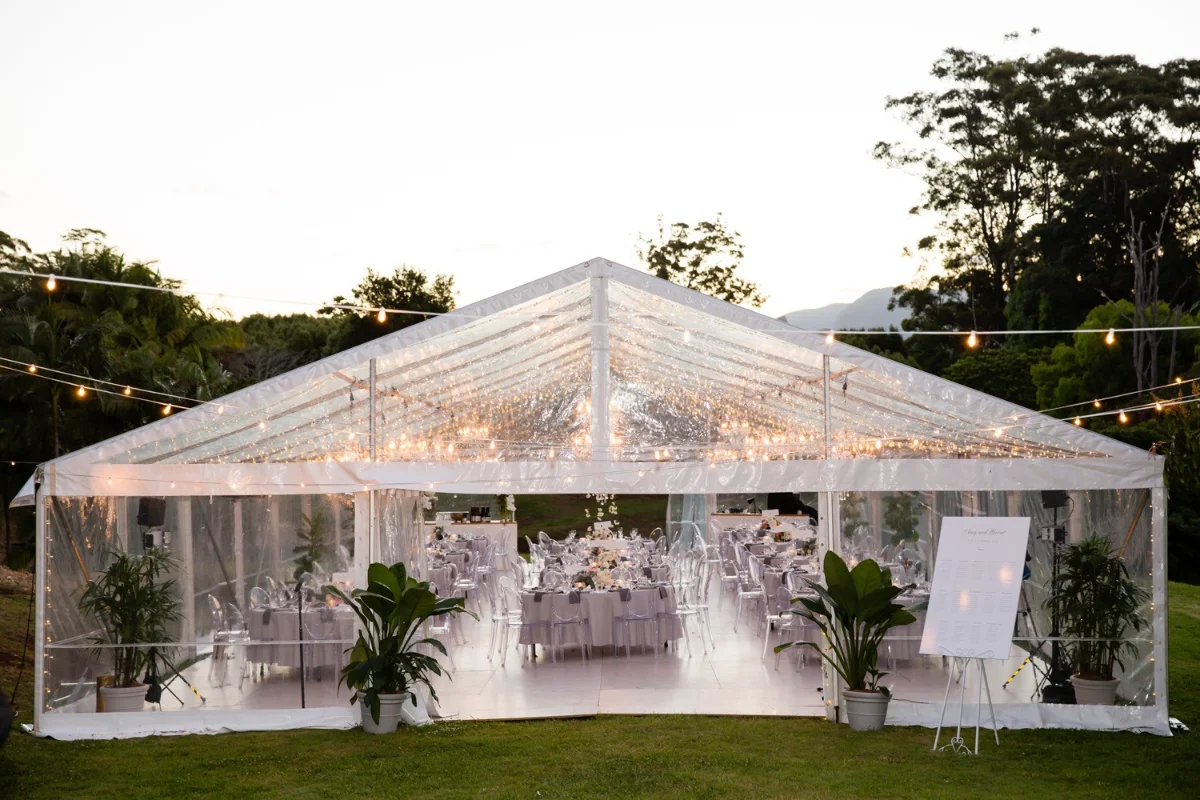 Clear-roof marquee glowing with fairy lights at dusk, set with round tables and ghost chairs, a seating chart on an easel and potted plants at the entrance