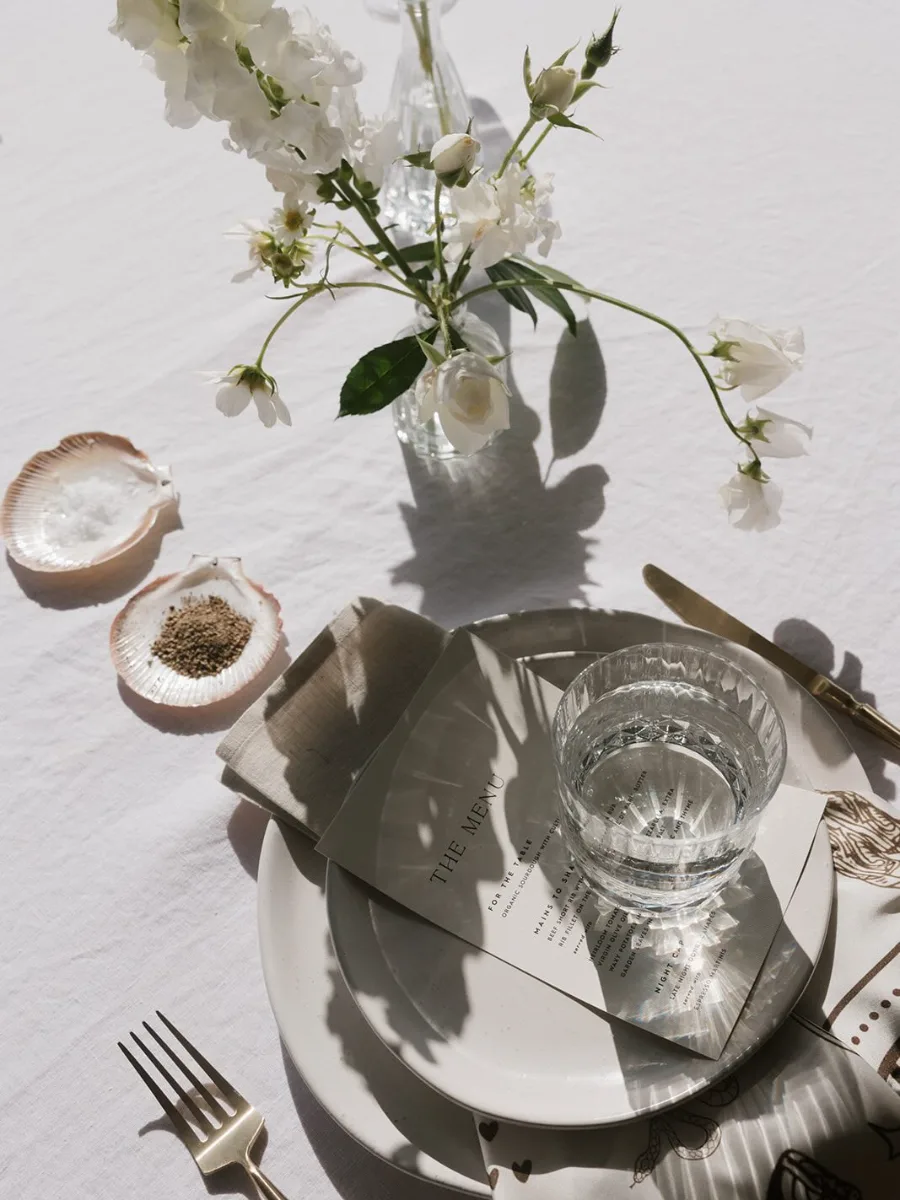 Overhead place setting on white linen with a printed menu, crystal tumbler, scallop-shell salt dishes and white sweet peas in a glass bud vase