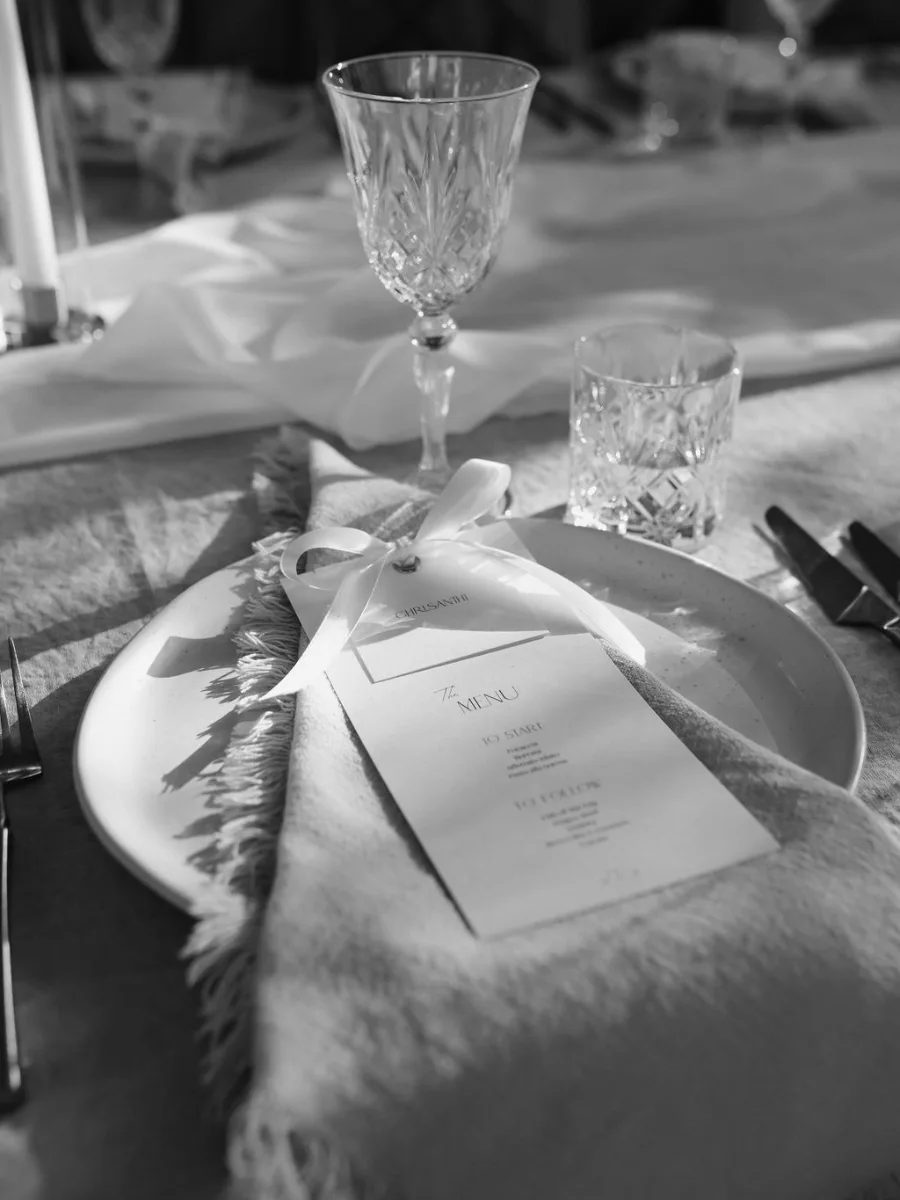 Black and white place setting with a ribboned menu card tucked into a fringed napkin, a crystal wine glass and tumbler