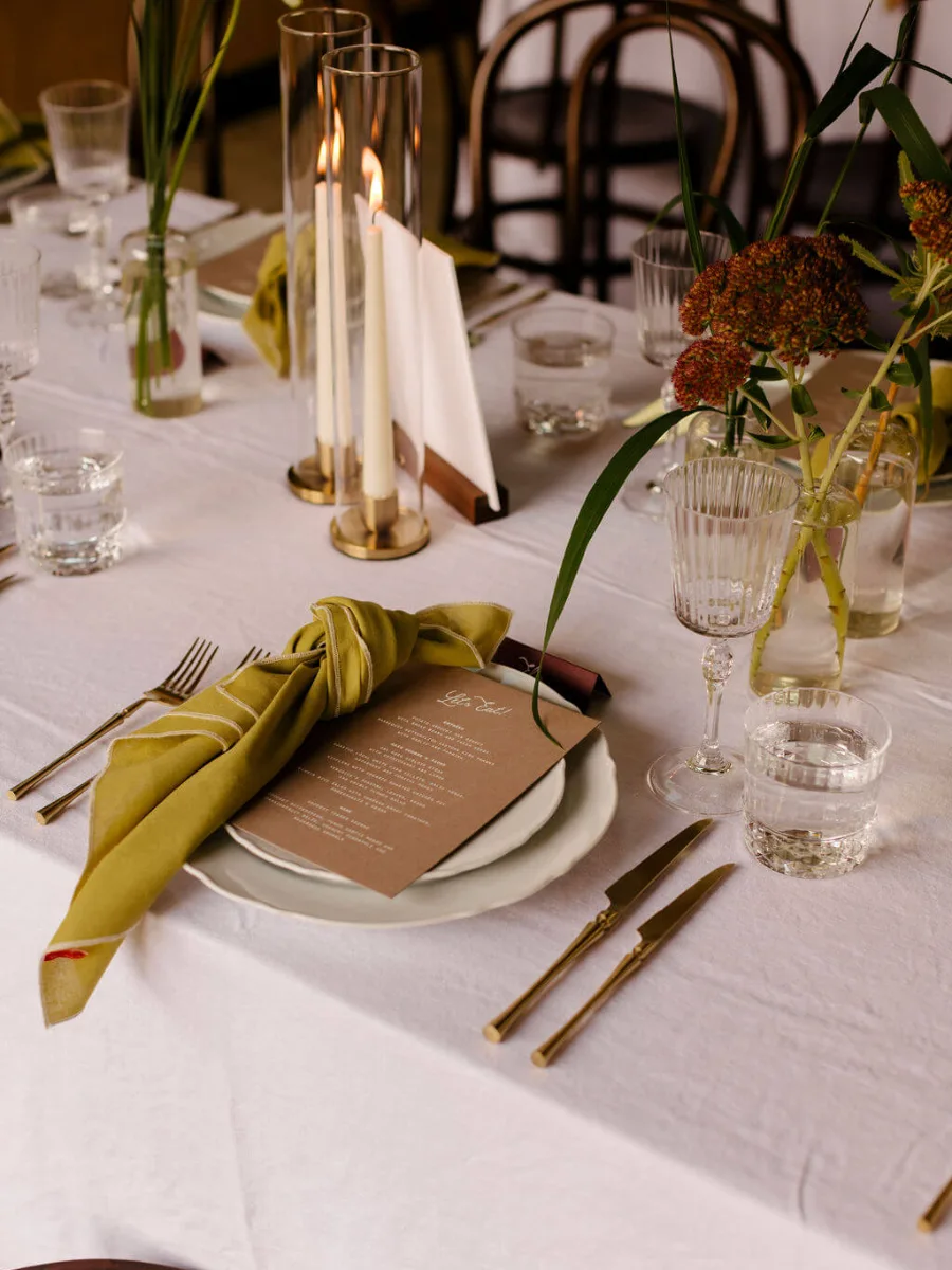 Place setting with an olive-green knotted napkin and kraft menu card, taper candles in glass hurricanes and rust-red sedum in bud vases on white linen