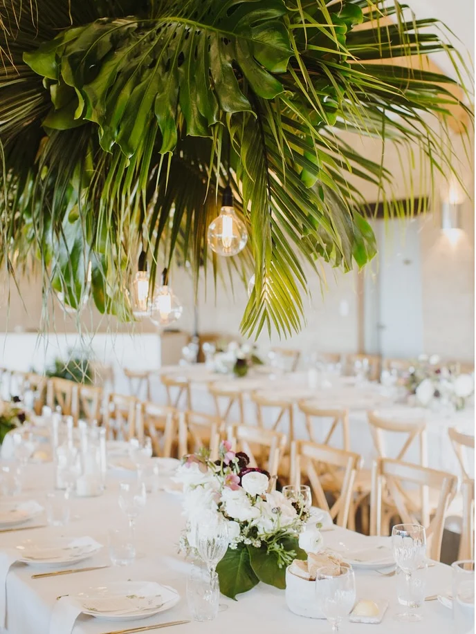 Hanging palm-frond canopy with pendant bulbs above a long white-linen table with a white floral centrepiece and cross-back chairs