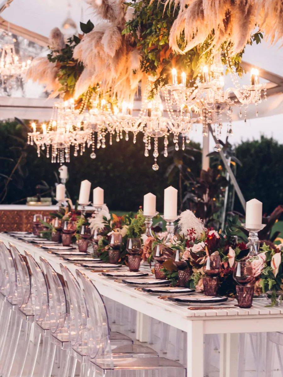 Long white table with pillar candles and burgundy glassware beneath hanging pampas grass and crystal chandeliers, with clear ghost chairs
