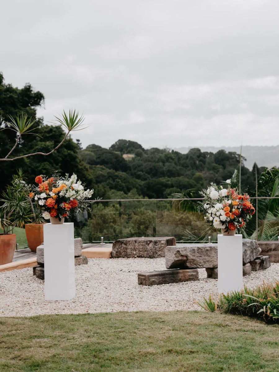 Two white pedestals holding orange, red and white floral arrangements on a gravel terrace with stone seating, overlooking the hinterland