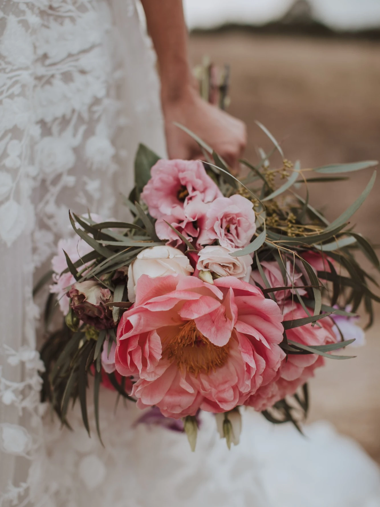 Bride in a floral lace gown holding a loose bouquet of coral peony, pink lisianthus and eucalyptus in a golden field