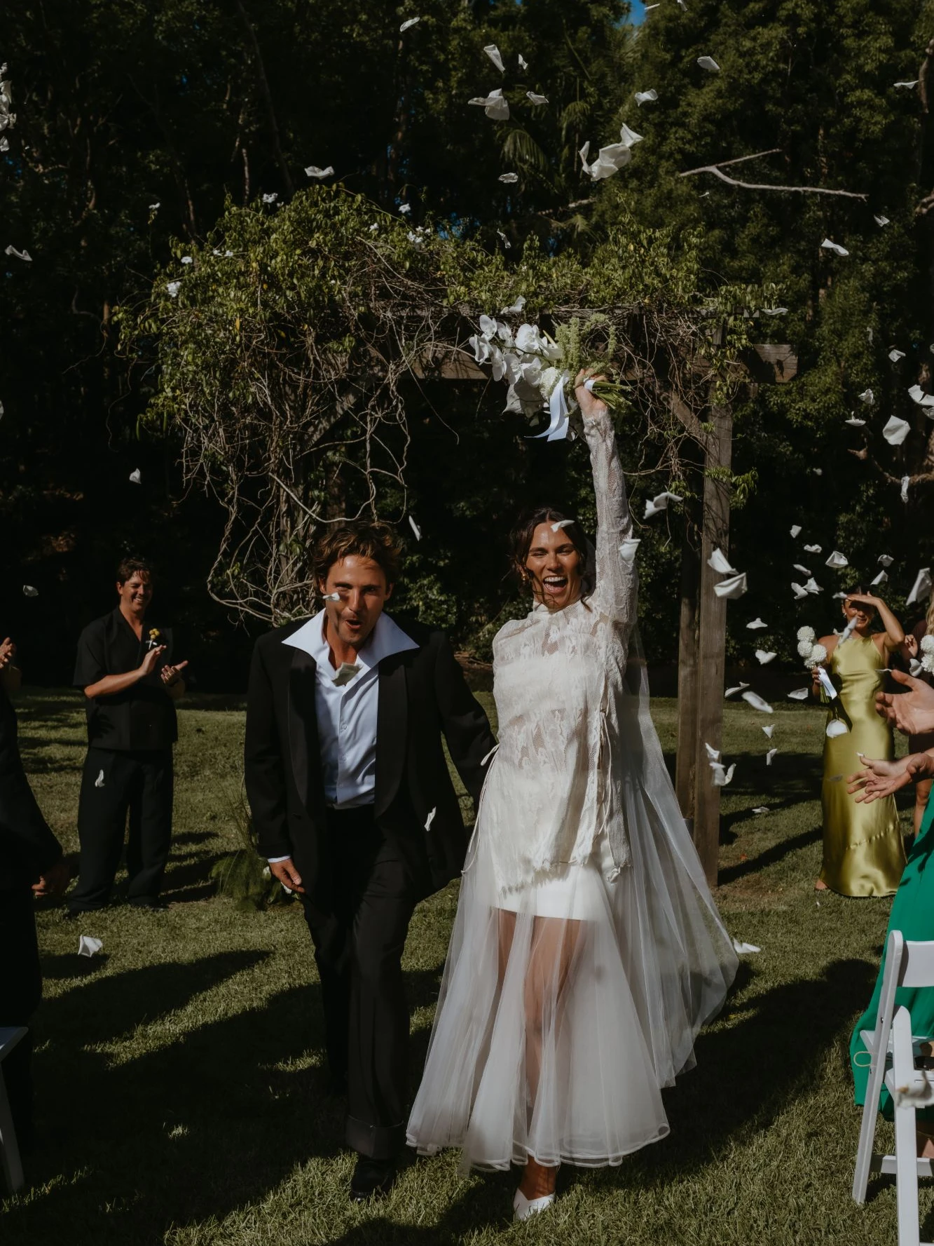 Laughing bride in a long-sleeved lace top and sheer tulle skirt raising her white bouquet beside her groom as petals fly through the air beneath a vine-covered arbour