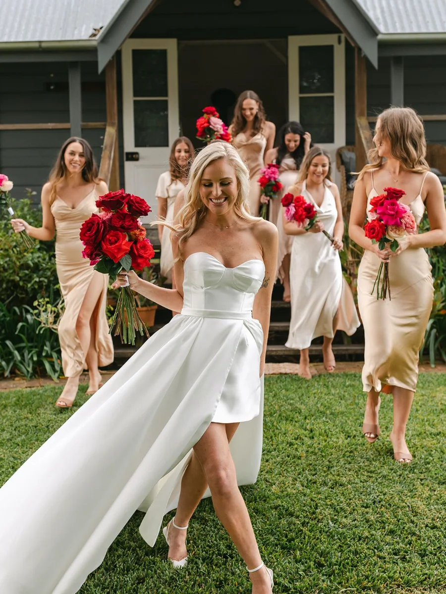 Laughing bride in a short satin dress with a long trailing skirt leading her bridesmaids, all holding red and pink rose bouquets, down the steps of a country cottage