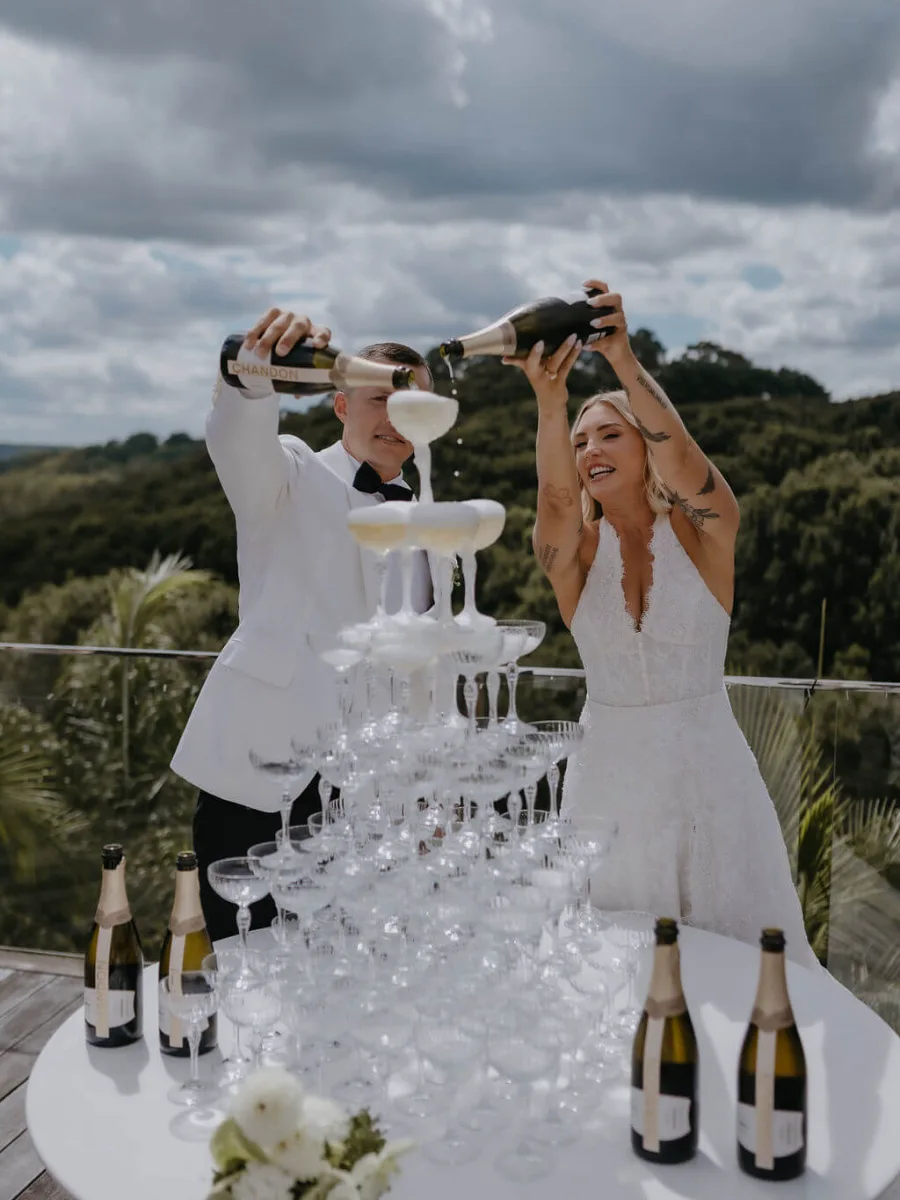Laughing couple pouring champagne from bottles over a coupe tower on a round white table, with bottles and a white bouquet, above forested hills