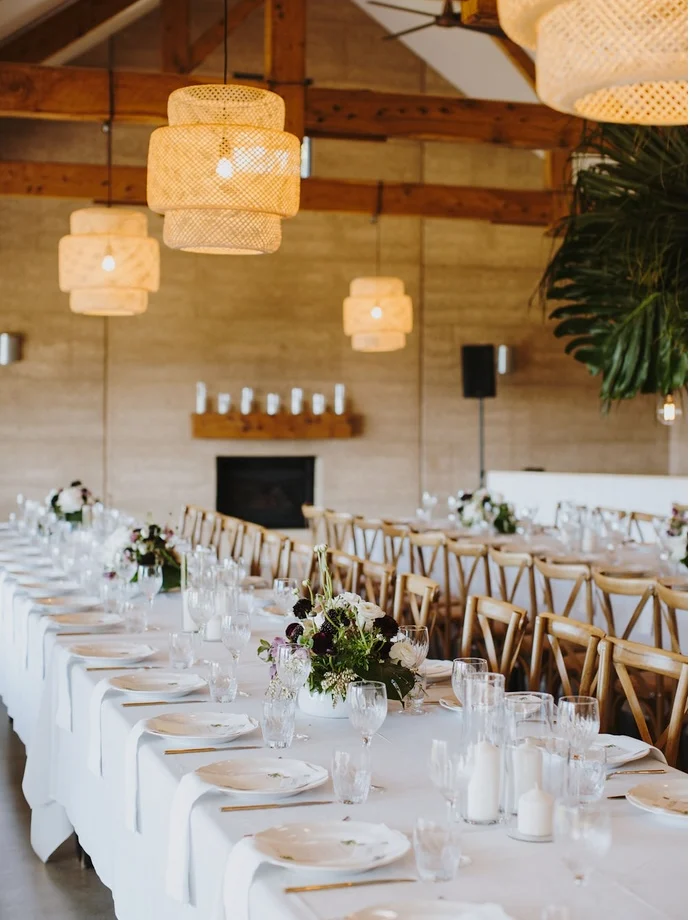 Long white-linen banquet table with cross-back chairs and a dark floral centrepiece beneath woven pendant lights in a timber-beamed barn