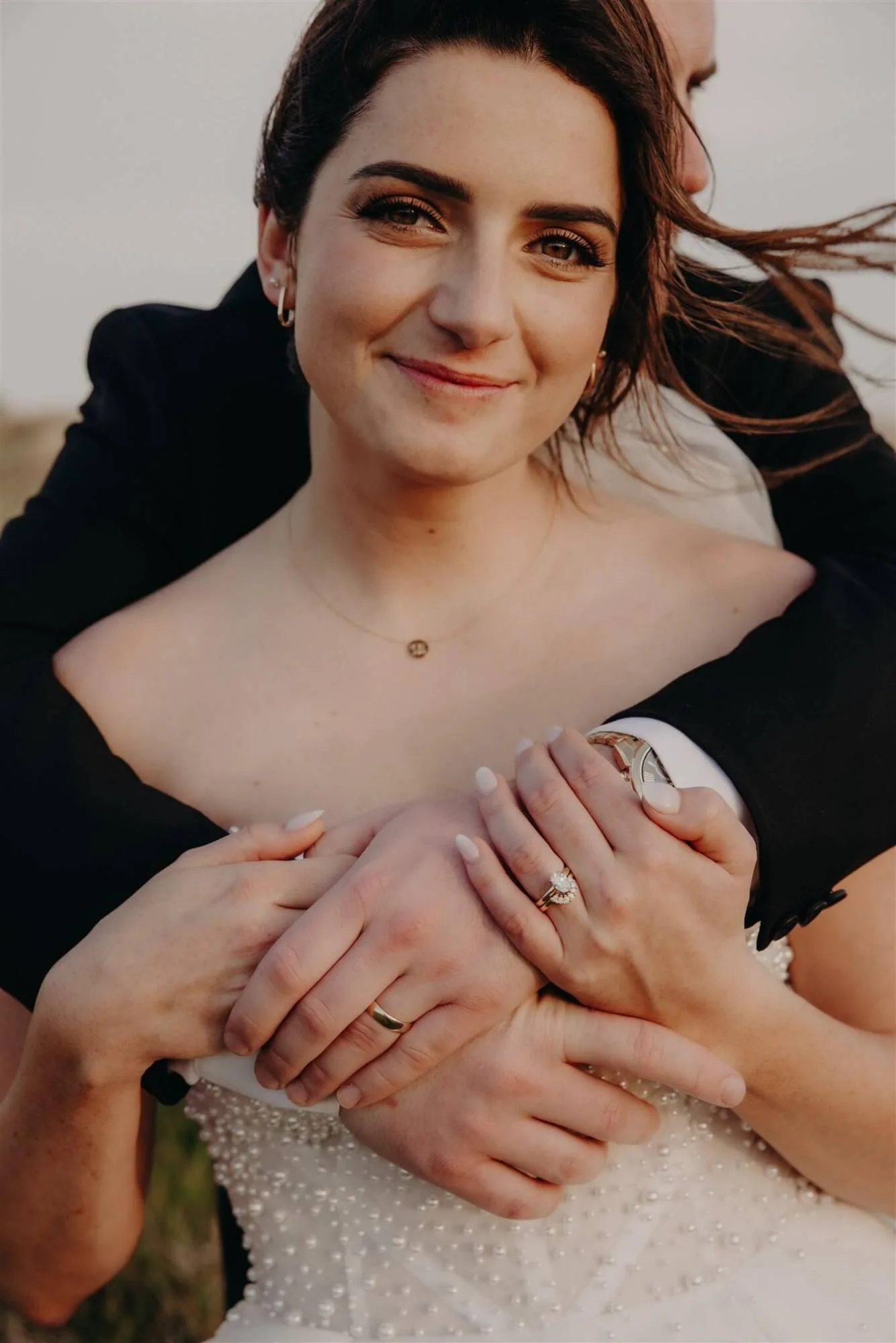 Close portrait of a smiling bride with windswept hair, her groom’s arms wrapped around her and both hands showing their rings