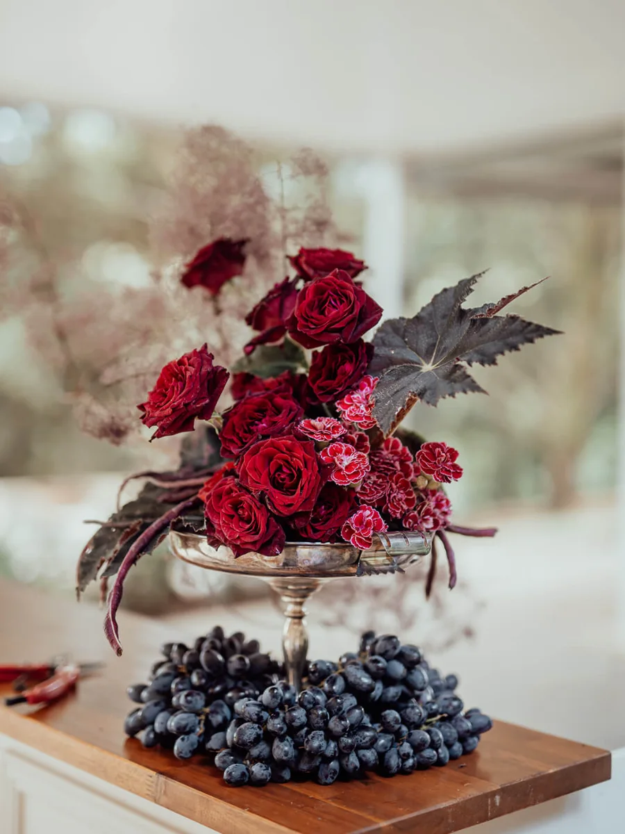 Silver compote bowl of deep red roses, pink carnations and a dark begonia leaf above a bunch of black grapes on a timber board