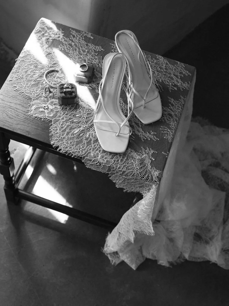 Black and white overhead of strappy bridal sandals, rings and jewellery resting on a lace-covered stool