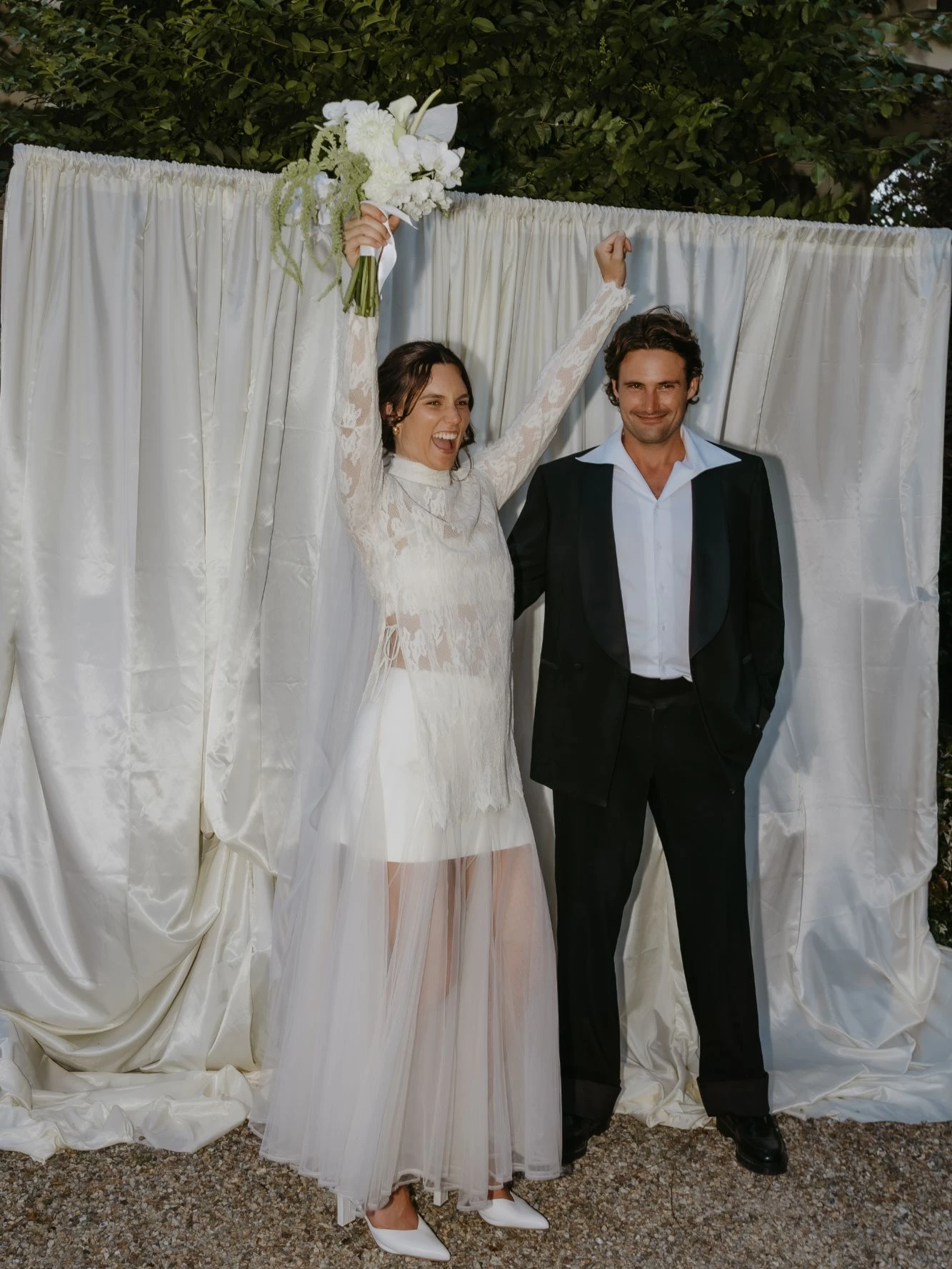 Bride in a lace top and sheer skirt raising her bouquet beside her groom in a black dinner jacket, in front of white satin drapes