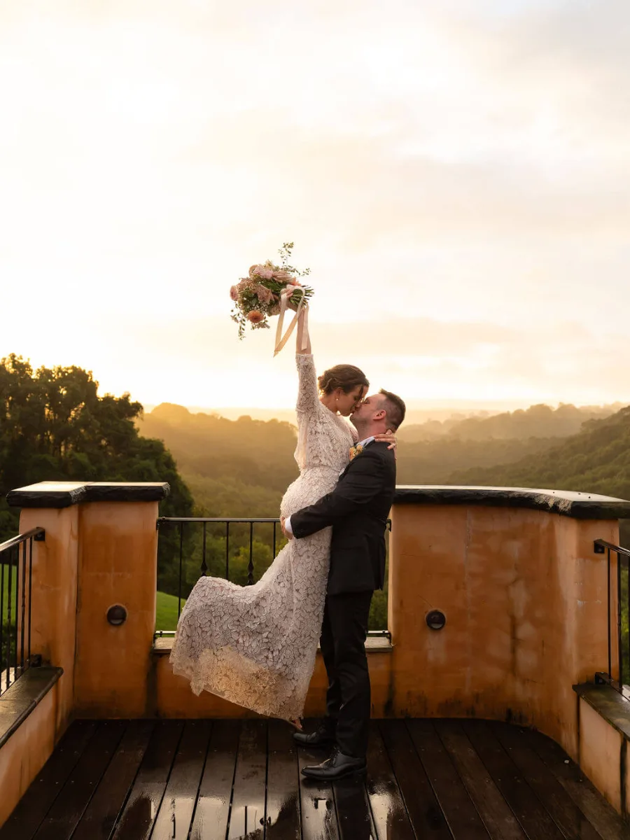 Groom lifting his bride in a kiss as she raises her bouquet at sunrise over the hinterland
