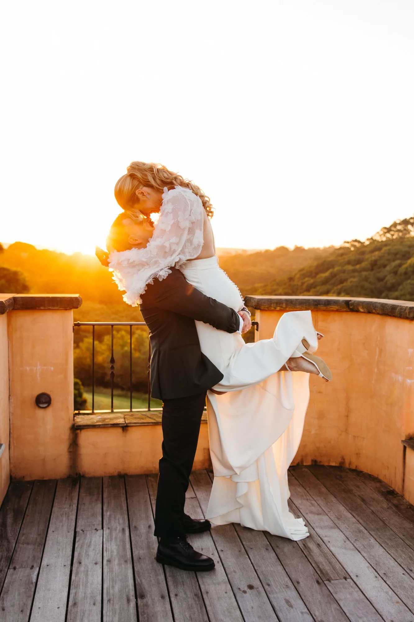 Groom lifting his bride in a kiss on a sunlit terrace, her gown and sleeves glowing in the golden hour with the hills behind