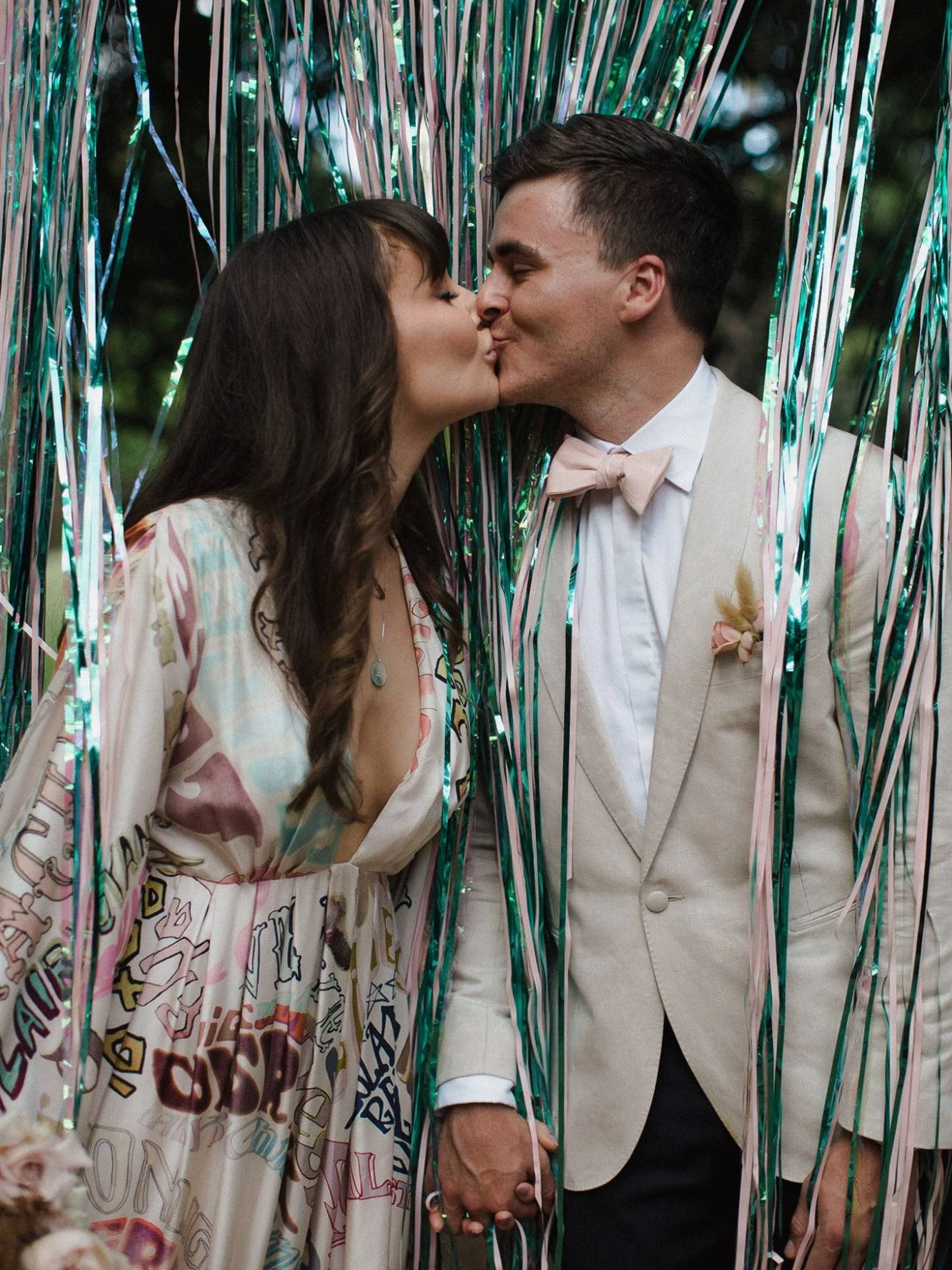 Bride in a hand-painted lettered robe kissing her groom in a cream jacket and pink bow tie, framed by green and pink tinsel curtains