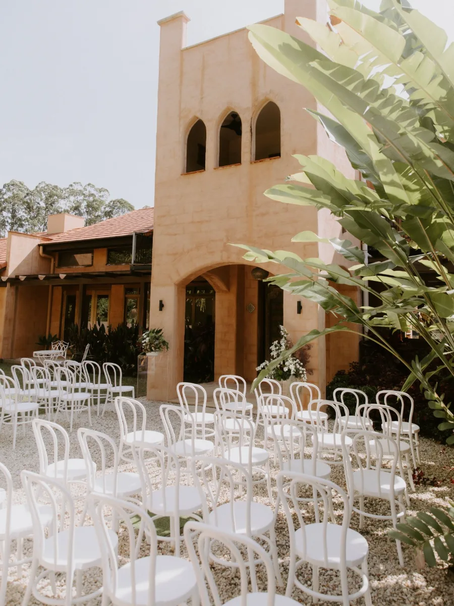 Rows of white bentwood chairs on a gravel courtyard facing a peach rendered tower with arched windows, white floral arrangements and banana palms