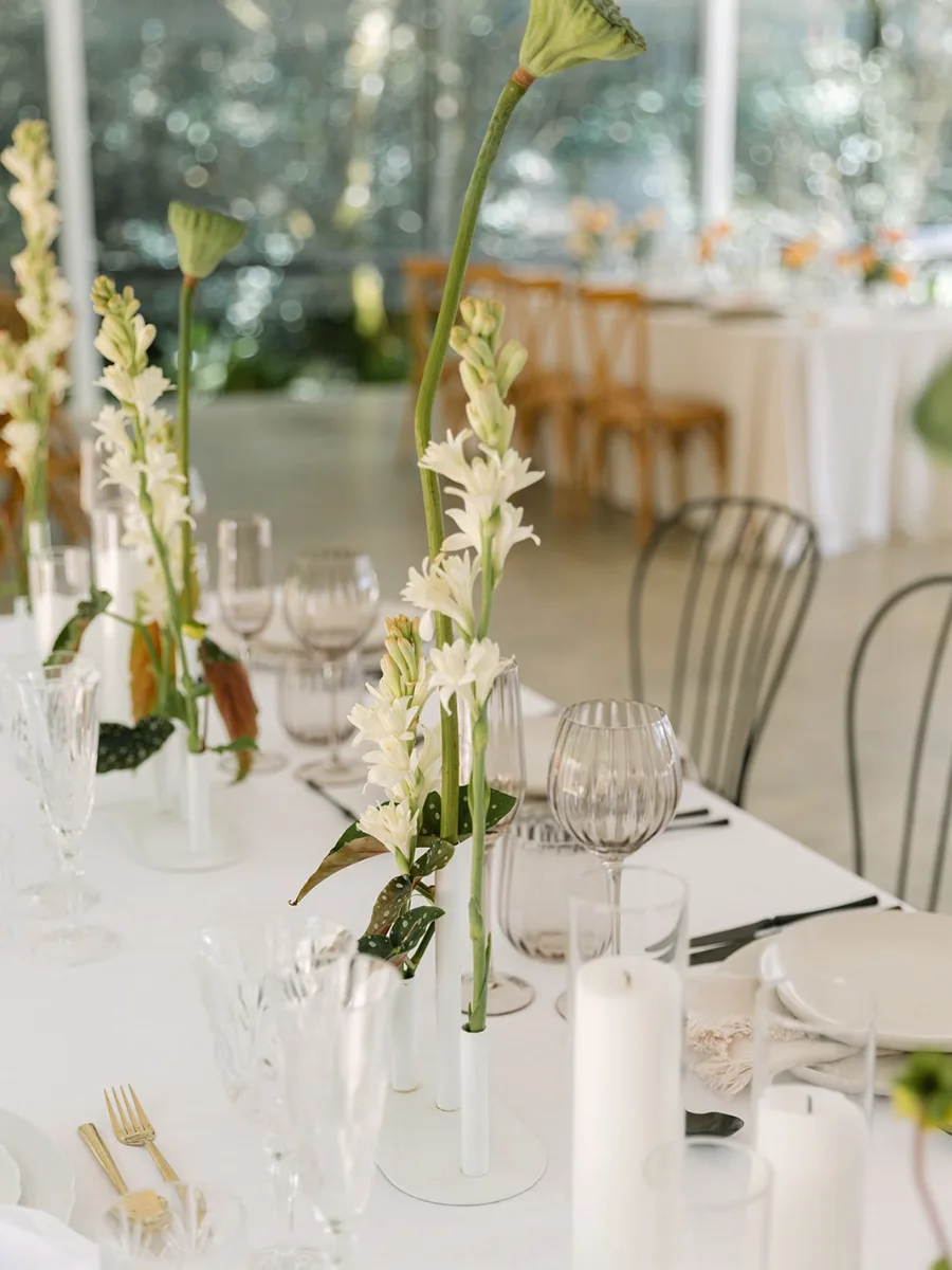 Tall tuberose stems and lotus seed pods in slim white vases on a white-linen table with crystal glassware, pillar candles and gold cutlery