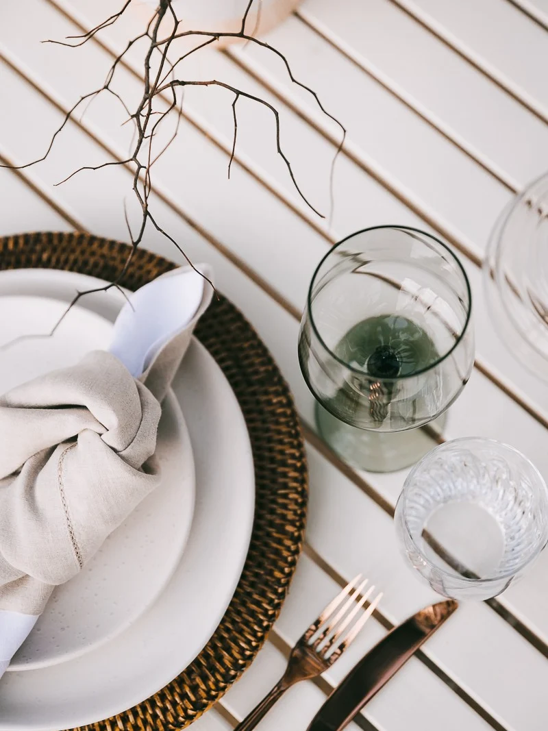 Overhead place setting with a knotted linen napkin on a woven rattan charger, a green wine glass, copper cutlery and a bare twig on a white slatted table