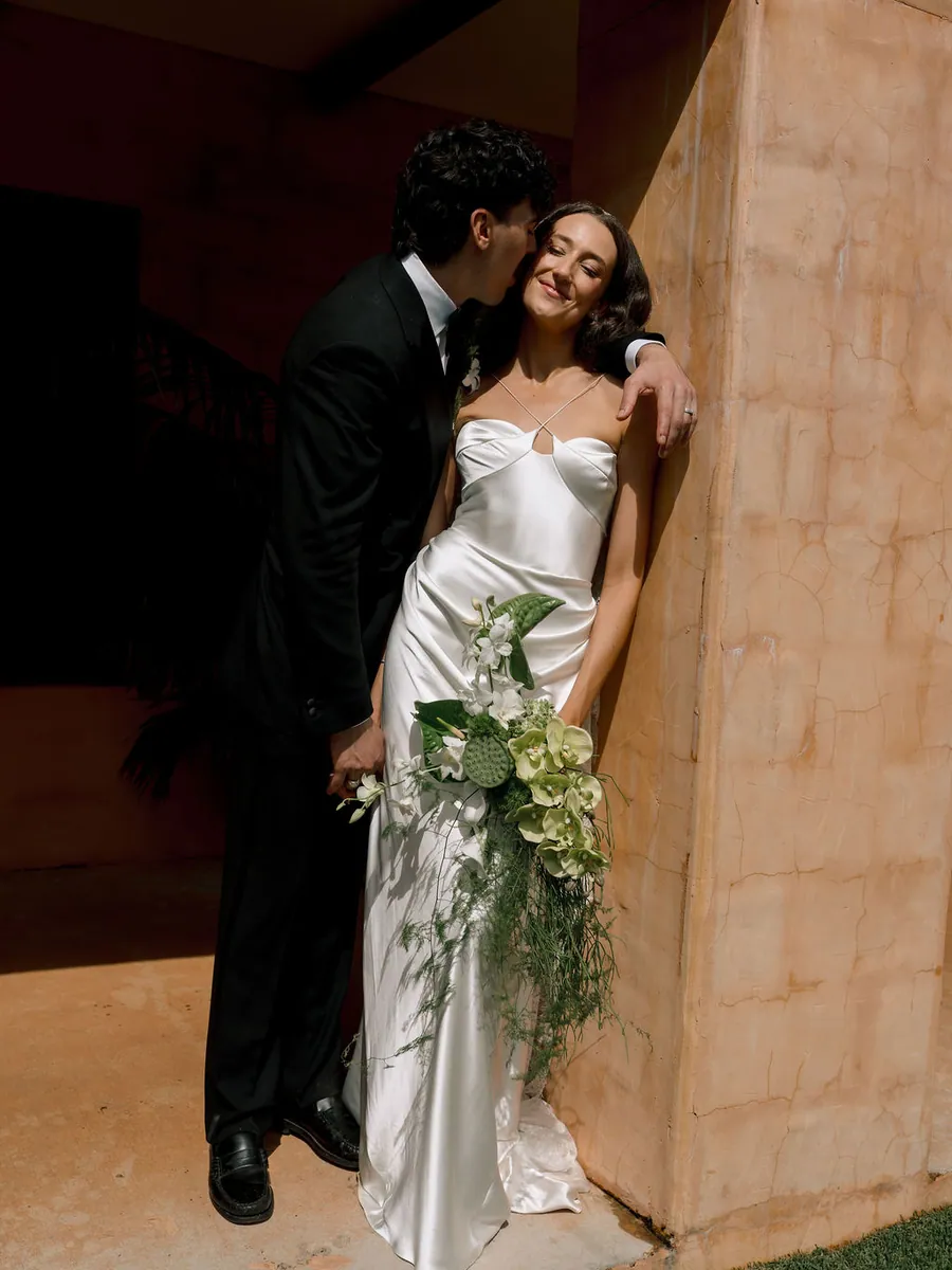 Groom kissing the bride beside a sandstone wall, her bouquet trailing in her hand