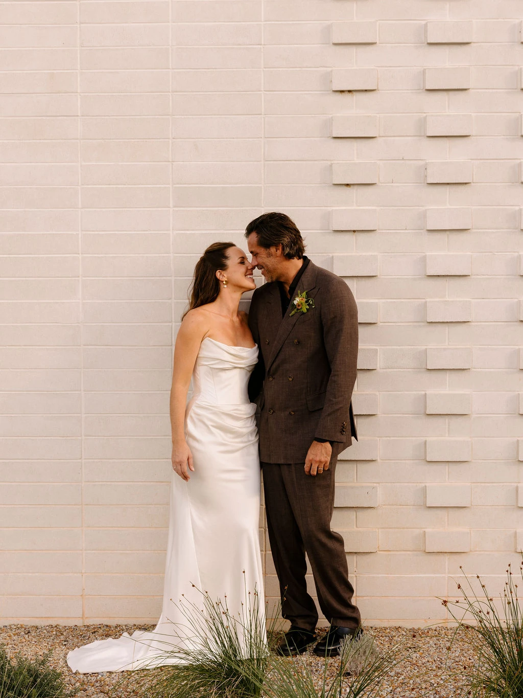 Smiling bride in a strapless satin gown touching noses with her groom in a brown double-breasted suit against a white patterned brick wall